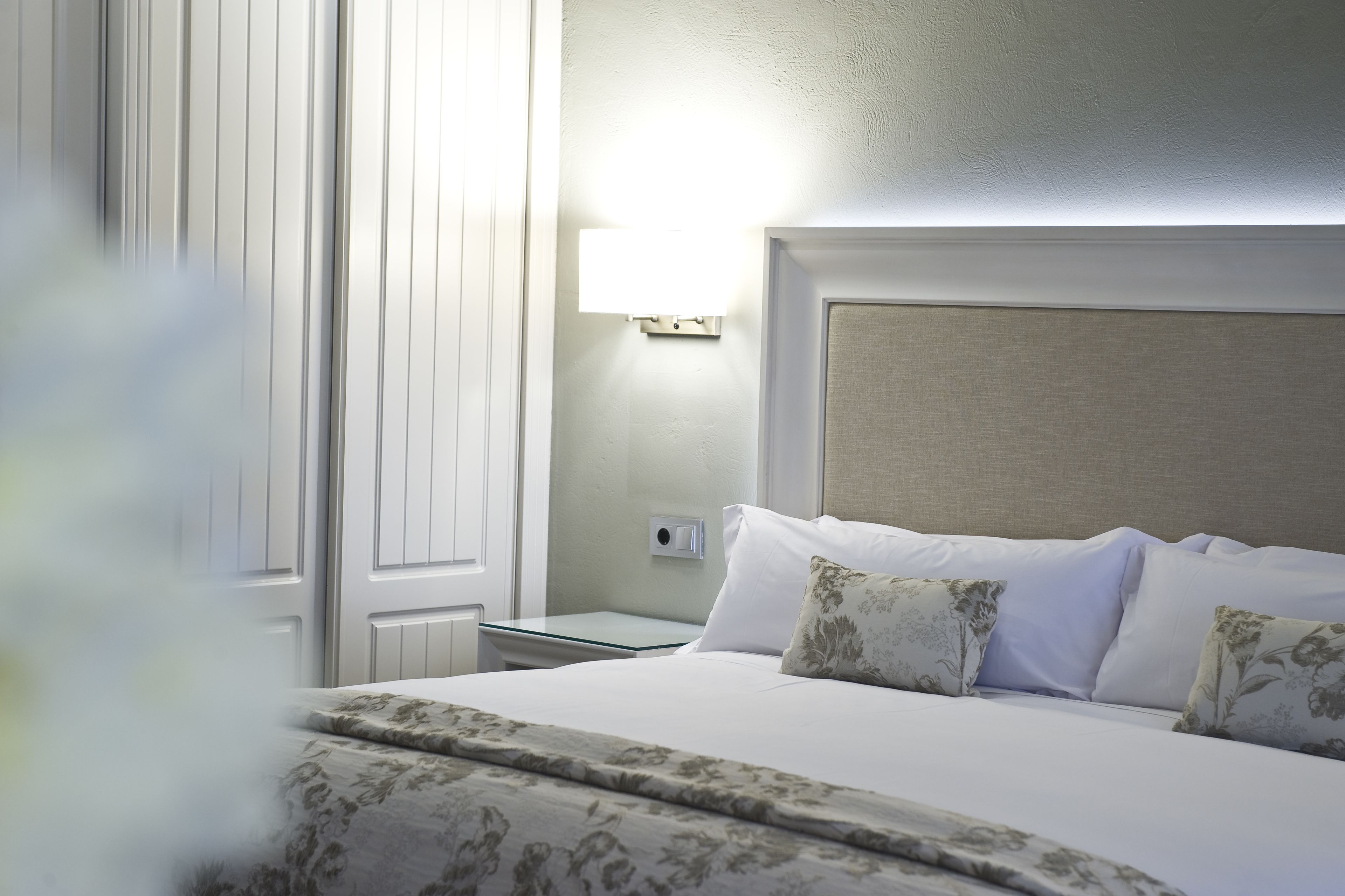 Una cama bien tendida con ropa de cama estampada con motivos florales, una mesita de noche con lámpara y una lámpara de pared.
