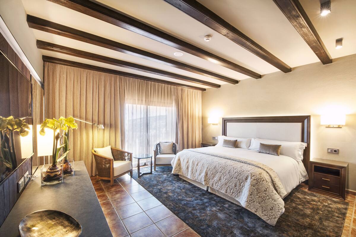Una habitación de hotel con una cama grande, un escritorio con silla, una mesita con un florero y un ventanal con cortinas.