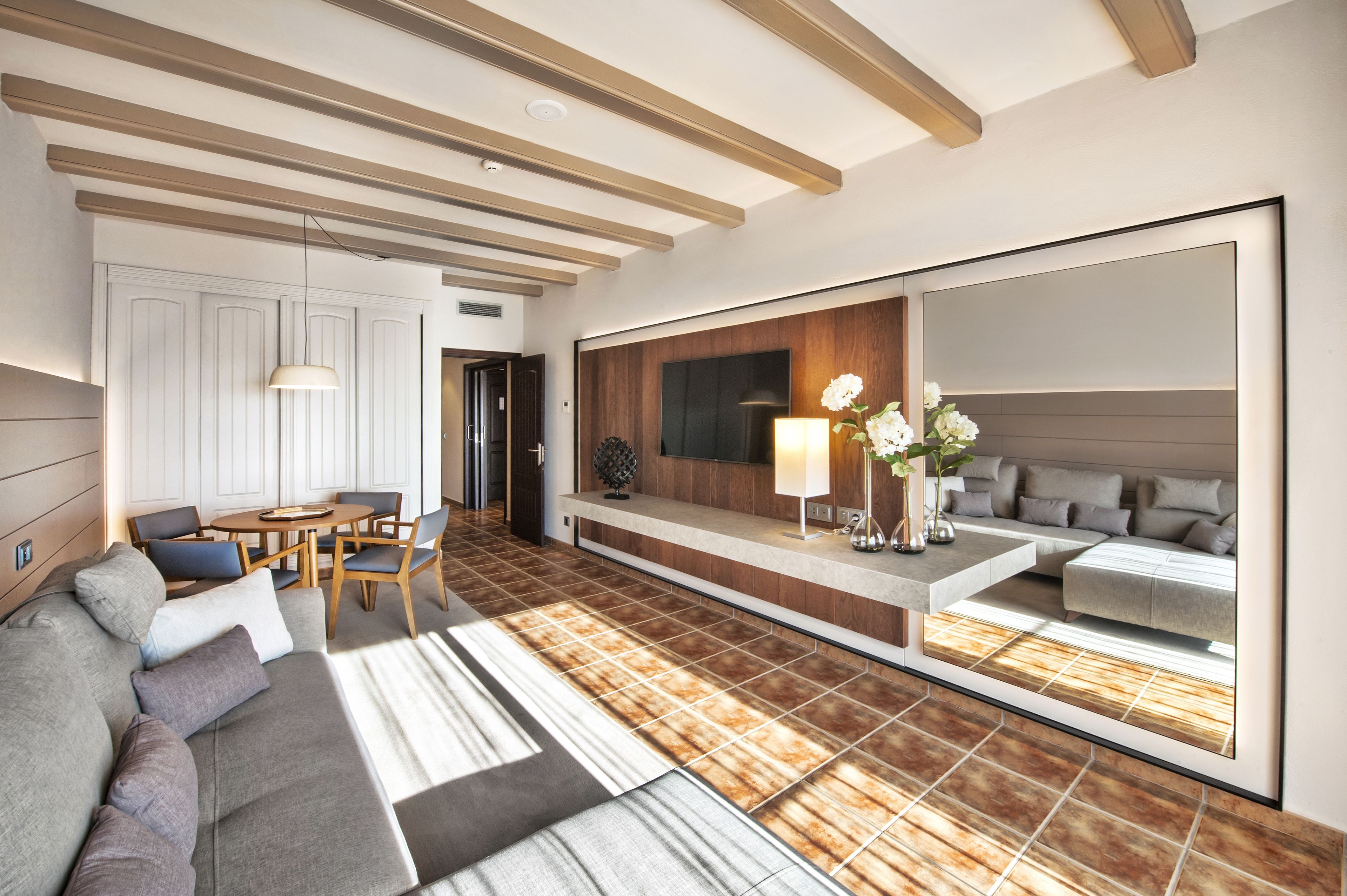 A modern living room with a sofa, dining table, chairs, and a large mirror reflecting a bedroom.