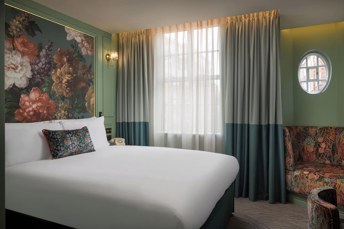 Een hotelkamer met een groot bed, een bloemmotief op de muur, een rond raam en een gestreept fauteuil.