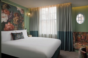 A hotel room with a large bed, a floral wall mural, a round window, and a patterned armchair.