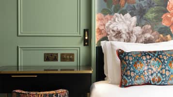 Een hotelkamer met een bed, een bureau en een stoel. De wand is bekleed met bloemenbehang en het bed heeft een kussen met een patroon.