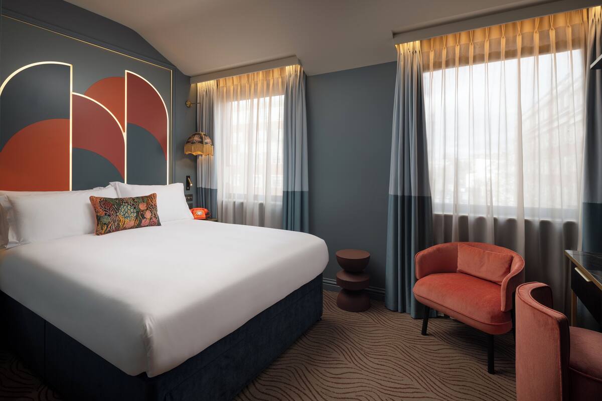 Een moderne hotelkamer met een groot bed, een zithoek met een stoel en uitzicht op de stad door grote ramen.