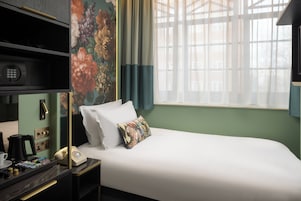 Ein Hotelzimmer mit einem Bett, einem Schreibtisch mit Telefon und Kaffeemaschine, einem Regal mit Radio und einem Fenster mit Vorhängen.