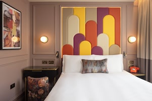 A hotel room with a large bed, a headboard with geometric patterns, a small table with a telephone, and a nightstand with a lamp.