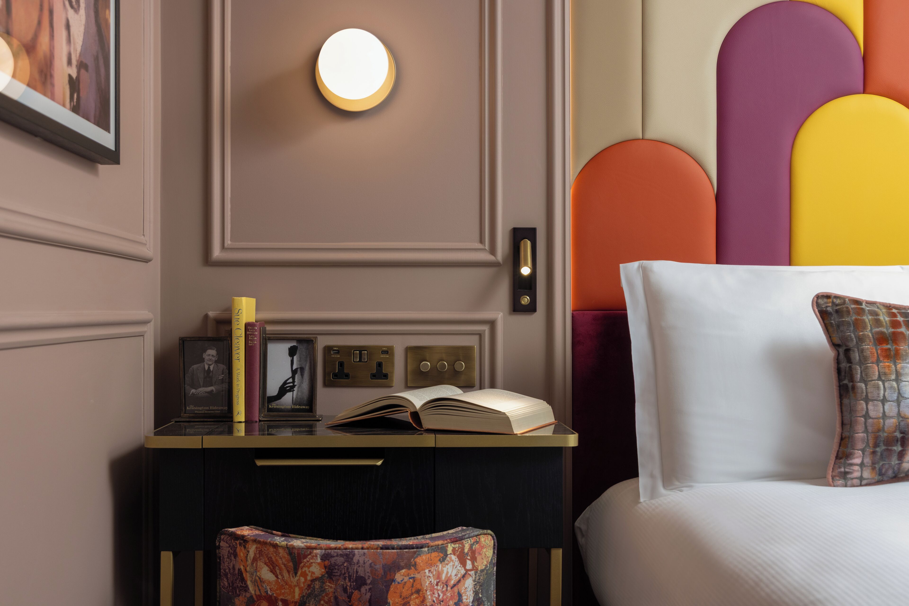 A bedroom with a bed, a bedside table with a book, a framed photo, and a wall-mounted light.