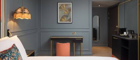 A hotel room with a bed, a desk, a chair, a lamp, a painting, a mirror, and a minibar.