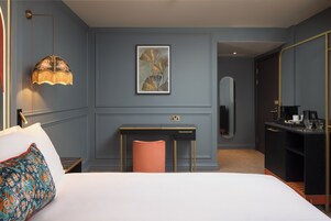 A hotel room with a bed, a desk, a chair, a lamp, a painting, a mirror, and a minibar.