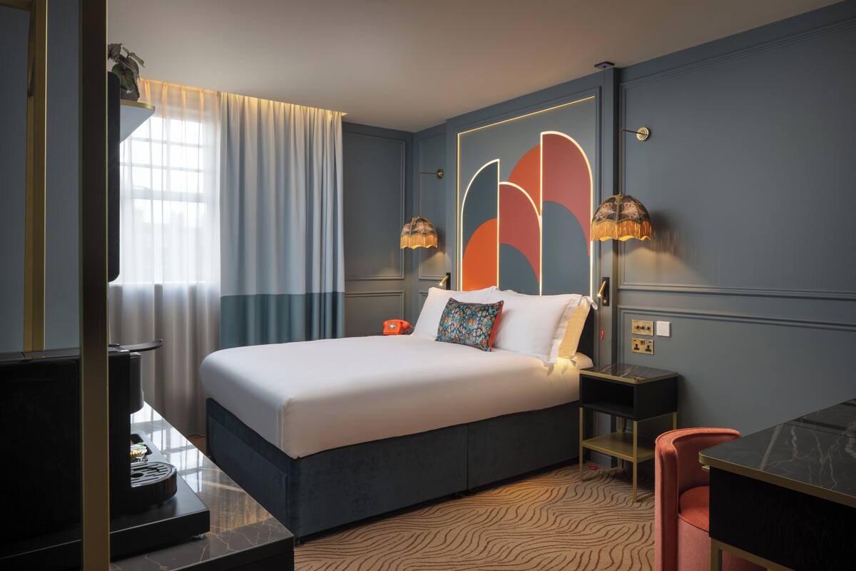 Een moderne hotelkamer met een groot bed, een bureau en een stoel. Er is een raam met gordijnen, een nachtkastje met een lamp, en een muur met een geometrisch ontwerp.
