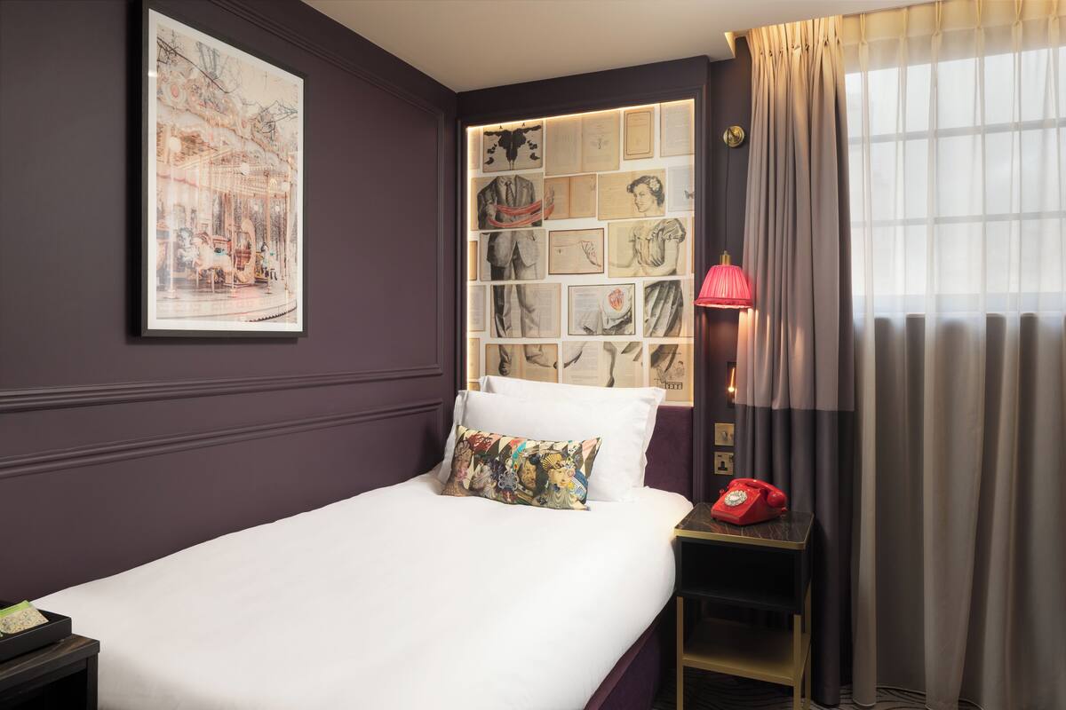 Een keurig opgemaakt bed met een kleurrijk kussen, een lijst met foto’s aan de muur, een rode draaischijf telefoon op een nachtkastje, en een raam met gordijnen.