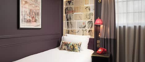 A neatly made bed with a colorful pillow, a framed collage of images on the wall, a red rotary phone on a bedside table, and a window with curtains.