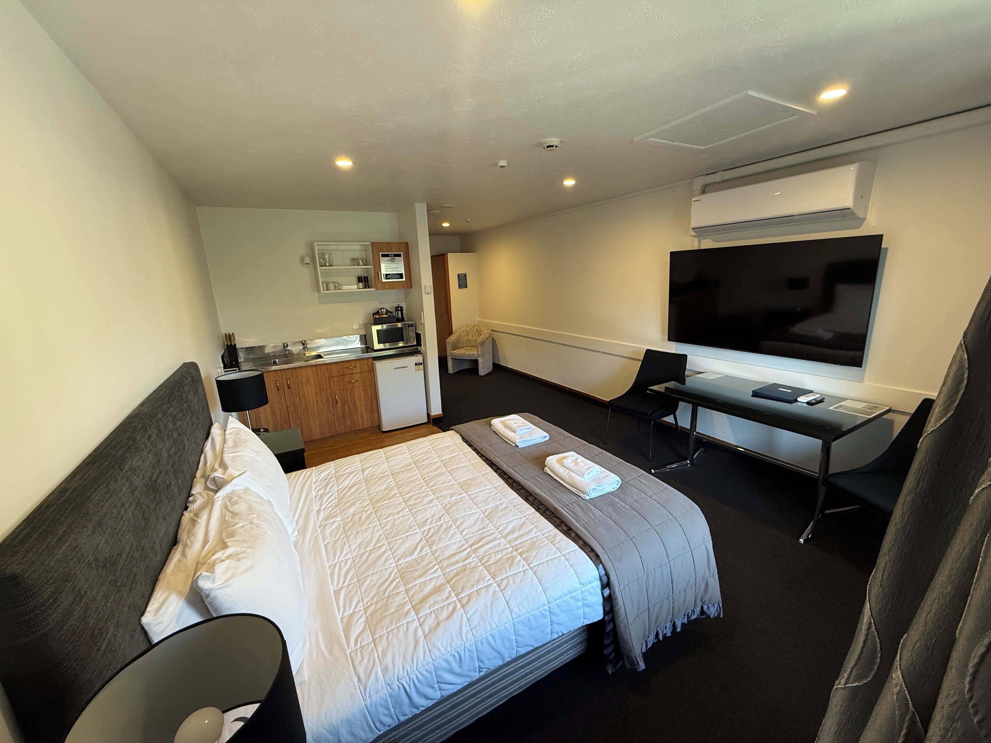 A hotel room with a bed, a desk, a chair, a TV, and a kitchenette.