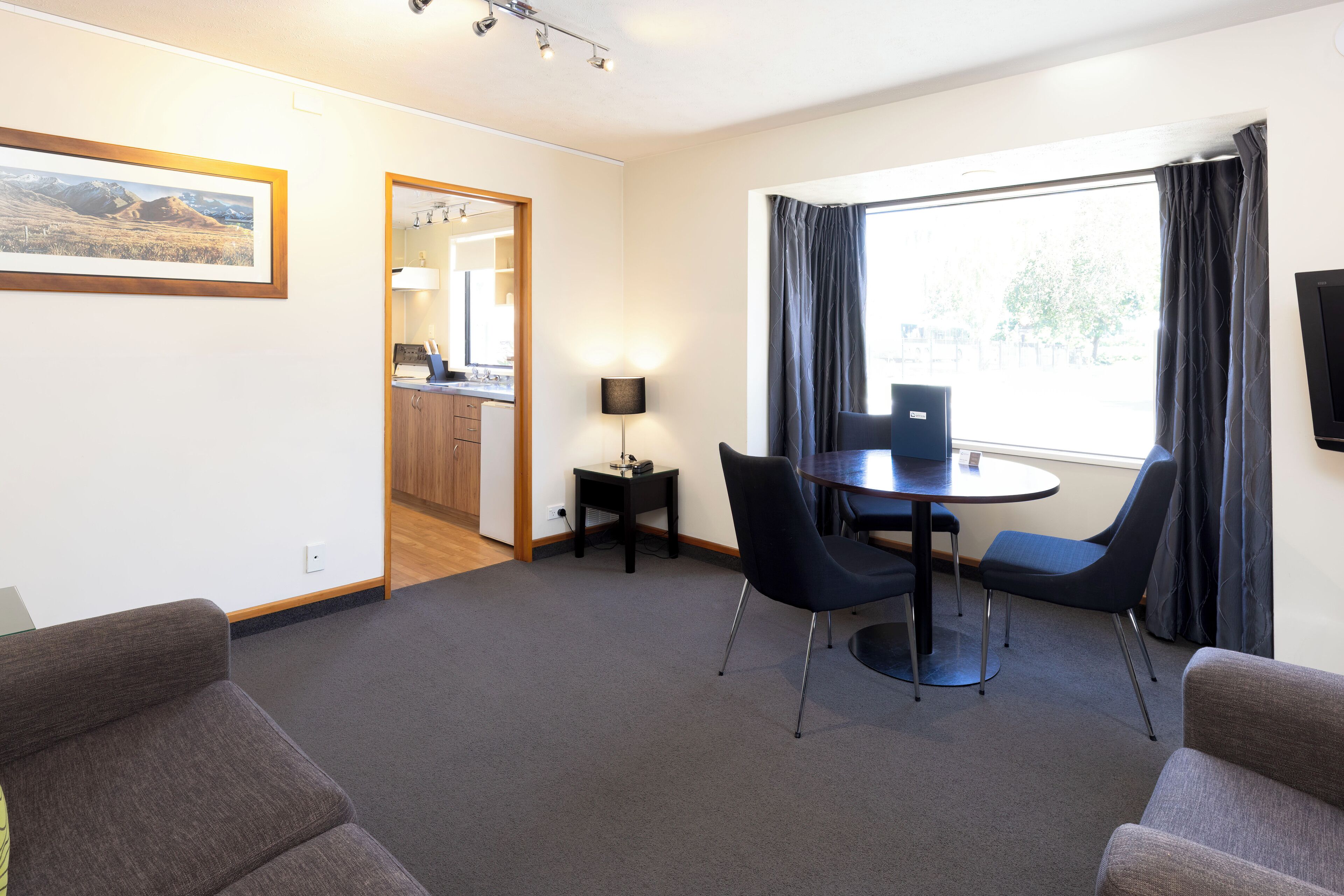 A modern hotel room with a dining area, a sofa, a small table with chairs, a lamp, and a TV on the wall.