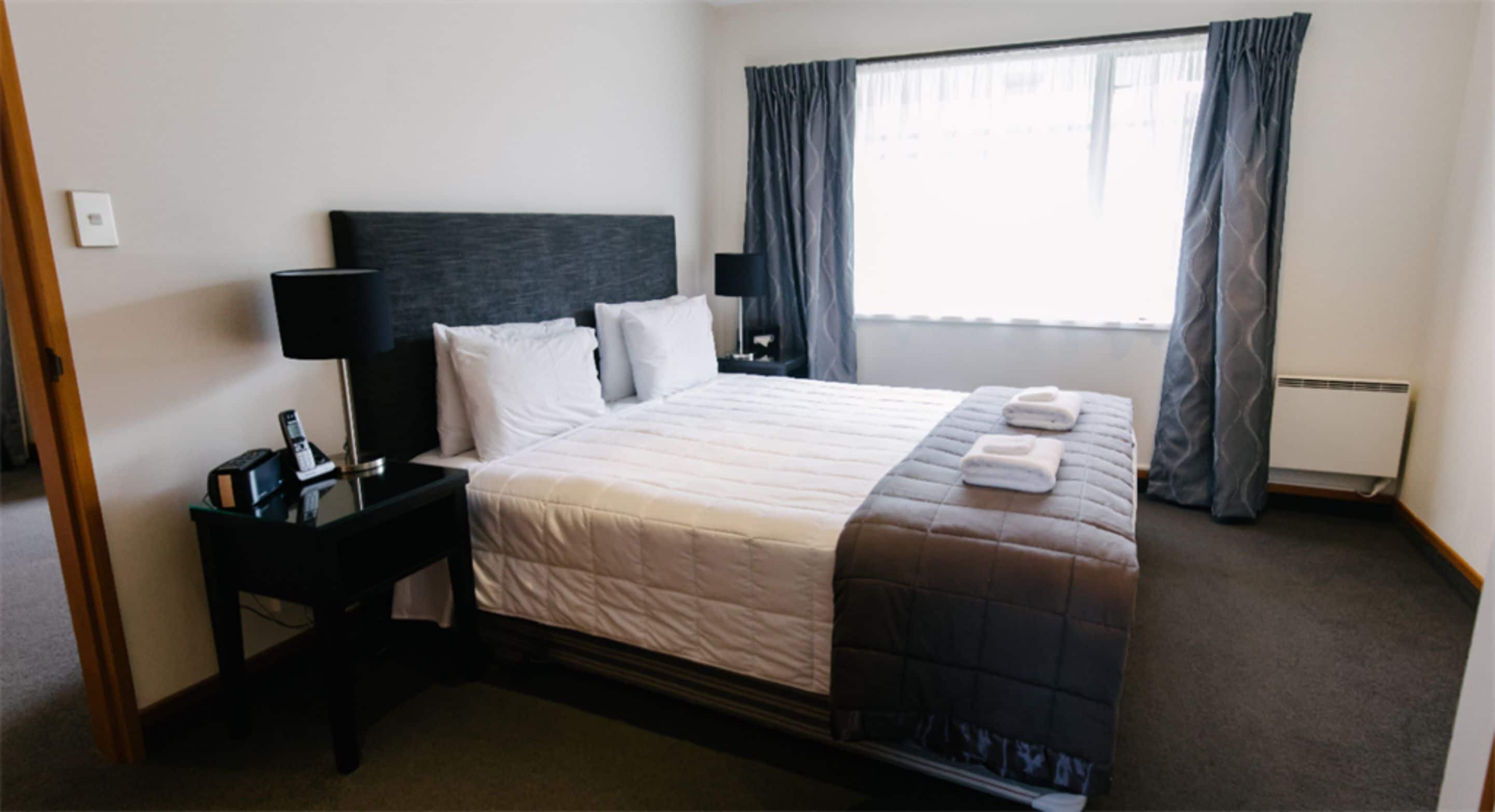 Quarto de hotel com uma cama grande, uma mesa de cabeceira com abajur, um criado-mudo e uma janela com cortinas.