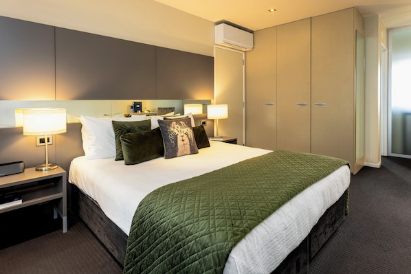A hotel room with a large bed, two bedside tables with lamps, a wardrobe, and an air conditioning unit.