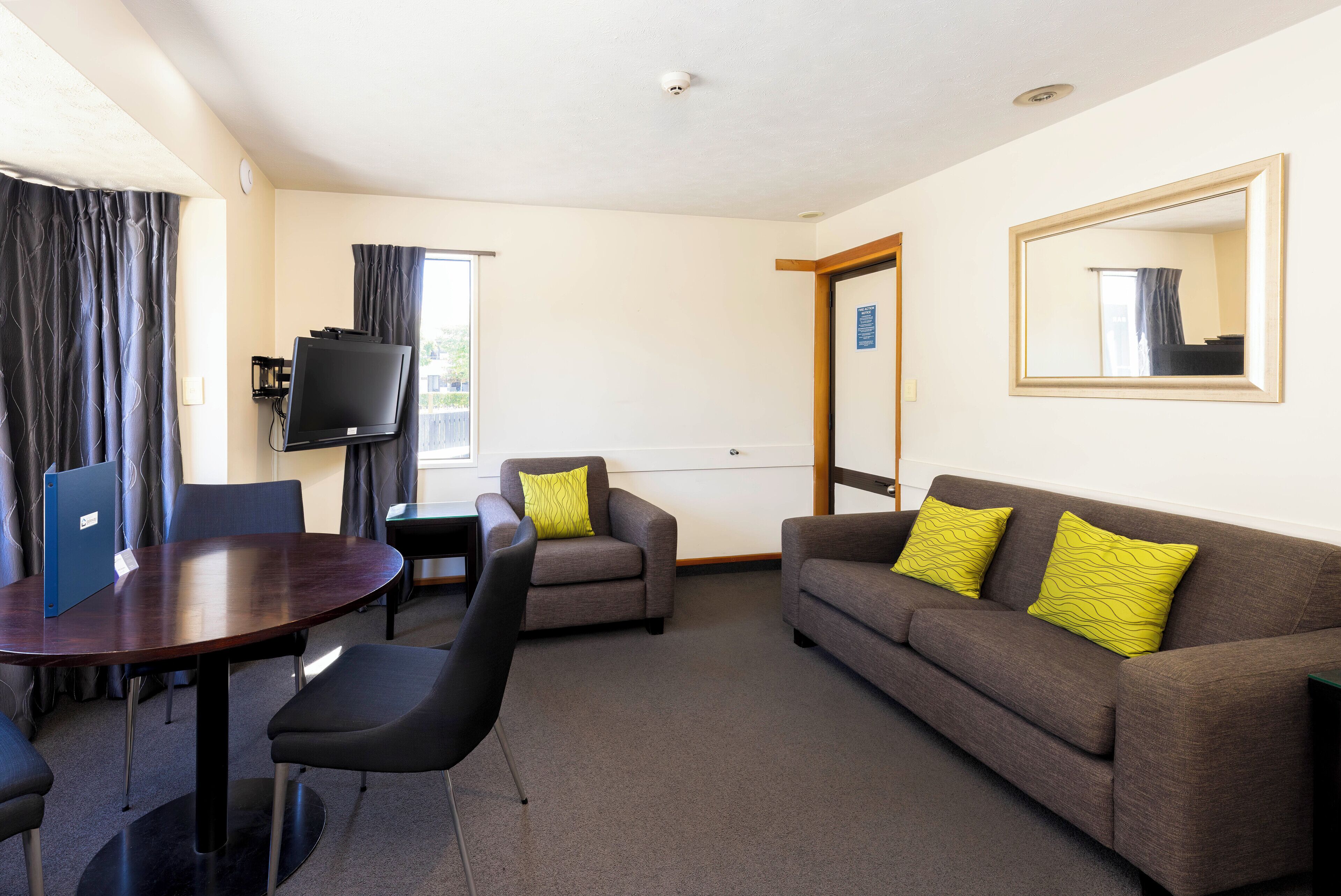 A hotel room with a round table, two armchairs, a sofa with yellow pillows, a TV, and a large mirror on the wall.