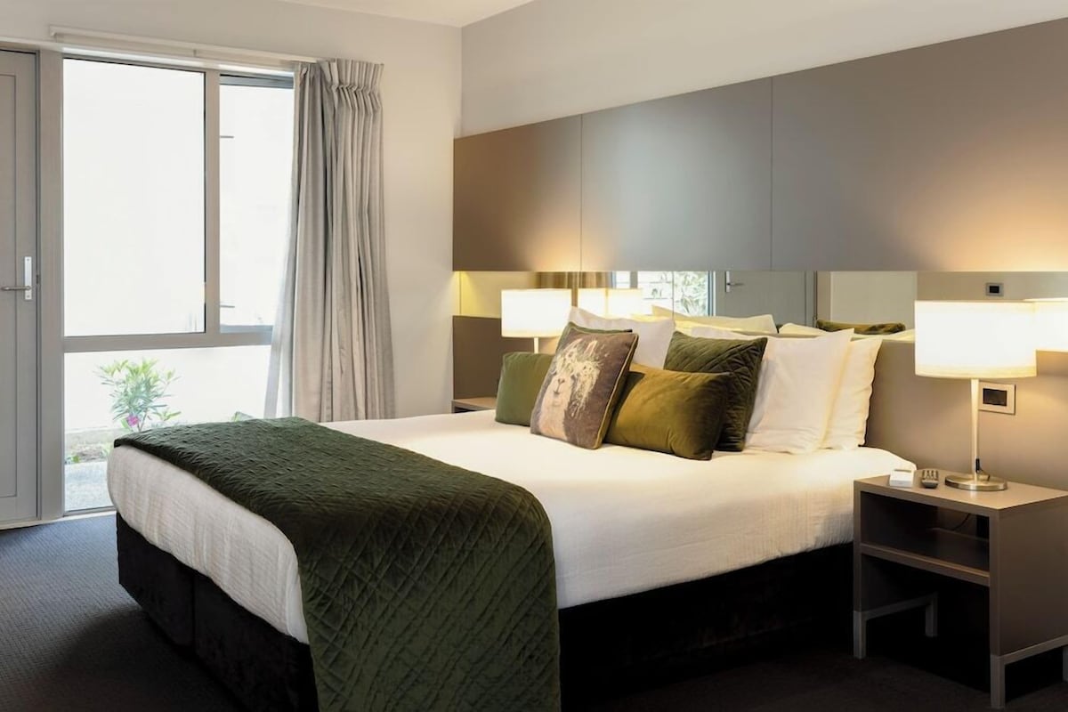 Een hotelkamer met een groot bed, twee nachtkastjes met lampen, een schuifdeur naar buiten en uitzicht op groen.