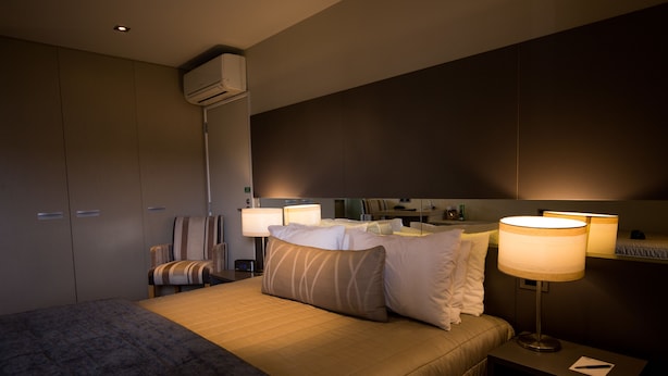 A hotel room with a large bed, two bedside tables with lamps, a chair, a wardrobe, and an air conditioning unit.