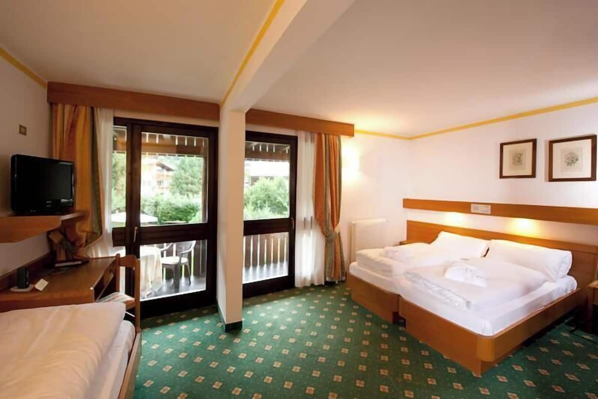 A hotel room with a bed, a desk, a television, and a balcony with a view of greenery.