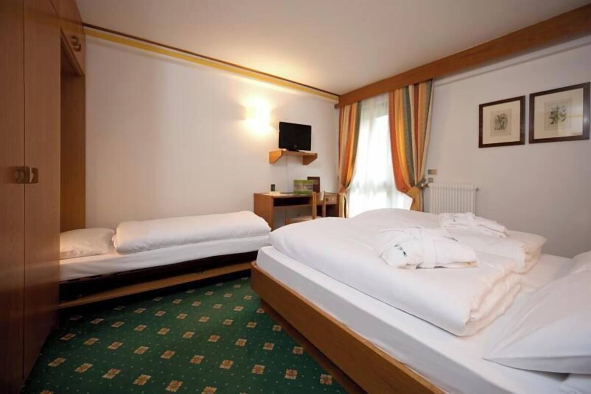 A hotel room with two single beds, a wooden wardrobe, a small desk with a chair, a television on a shelf, and a window with curtains.