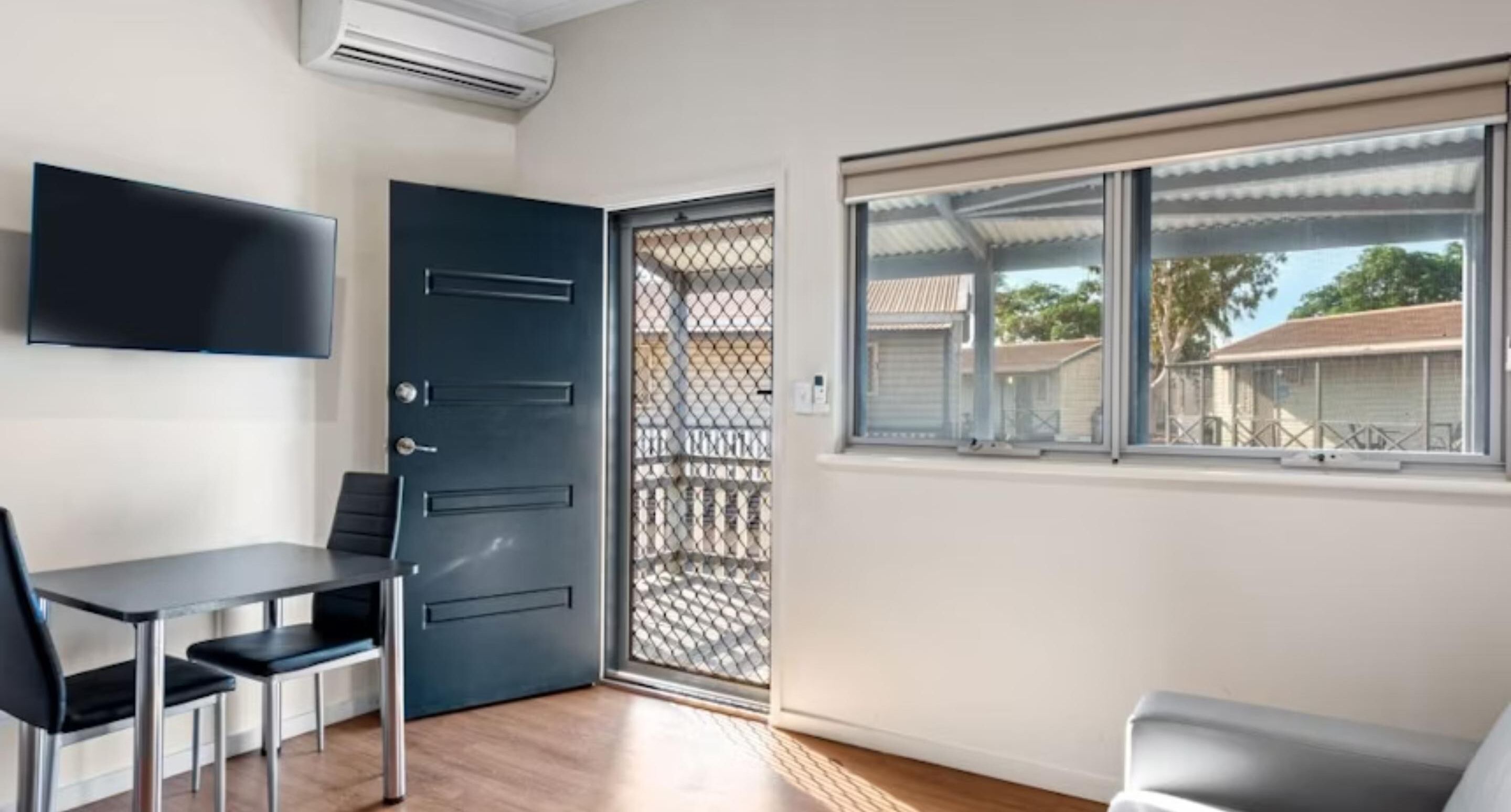 A room with a dark blue door, a TV mounted on the wall, a window with a view of houses, and a table with chairs.