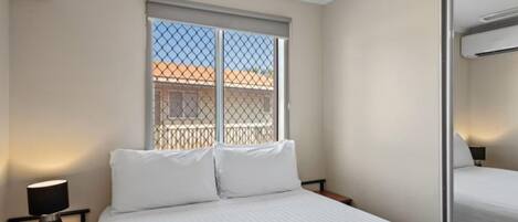 A hotel room with a bed, a nightstand with a lamp, and a window with a view of buildings.
