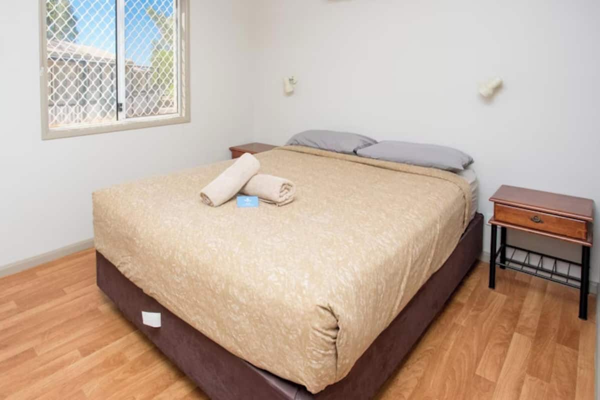 Ein ordentlich bezogenes Bett mit beiger Decke, ein kleiner Nachttisch mit Schublade und ein Fenster mit Metallfliegengitter.