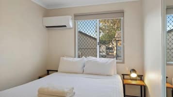 A bedroom with a bed, white linens, a nightstand with a lamp, an air conditioning unit, and a window with a view of buildings and trees.