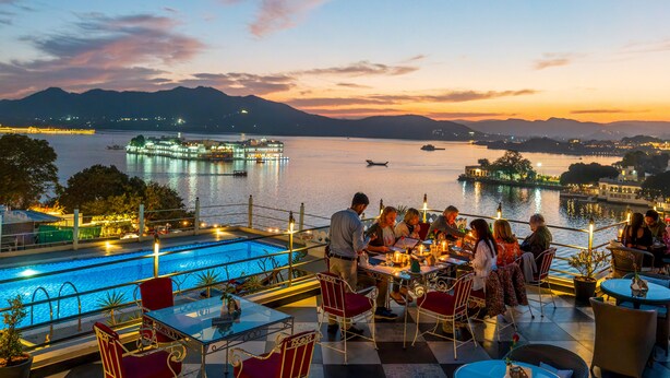 A rooftop bar with tables and chairs overlooking a lake, with mountains in the background. - Udaigarh (Udaipur)