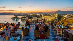 A terrace with tables and chairs, overlooking a body of water and a cityscape with illuminated buildings. - Udaigarh (Udaipur)