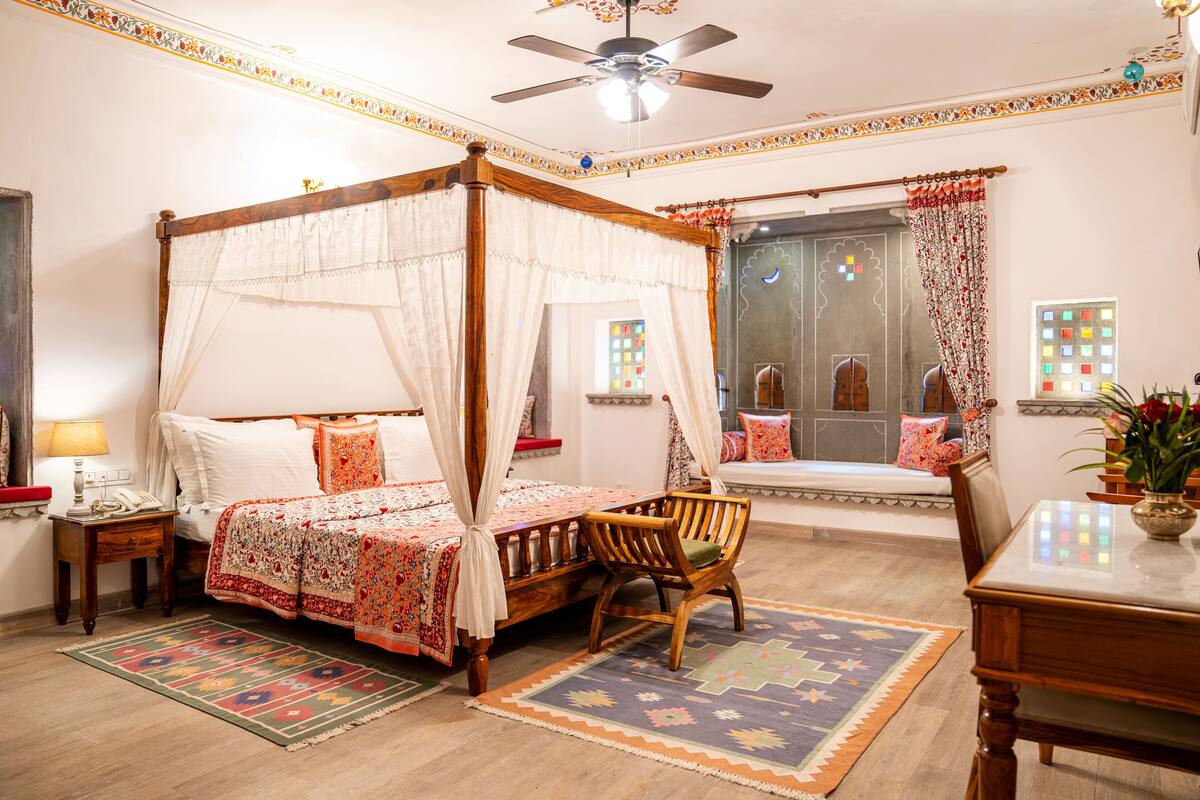 A four-poster bed with a canopy, a wooden chair, a small table, and a patterned rug in a room with a ceiling fan, a window with curtains, and a wall-mounted telephone.