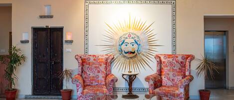A lobby with two patterned armchairs, a glass coffee table, and a large decorative wall piece.
