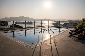 Piscina con vistas a una puesta de sol sobre montañas, barandilla y sillas de descanso.