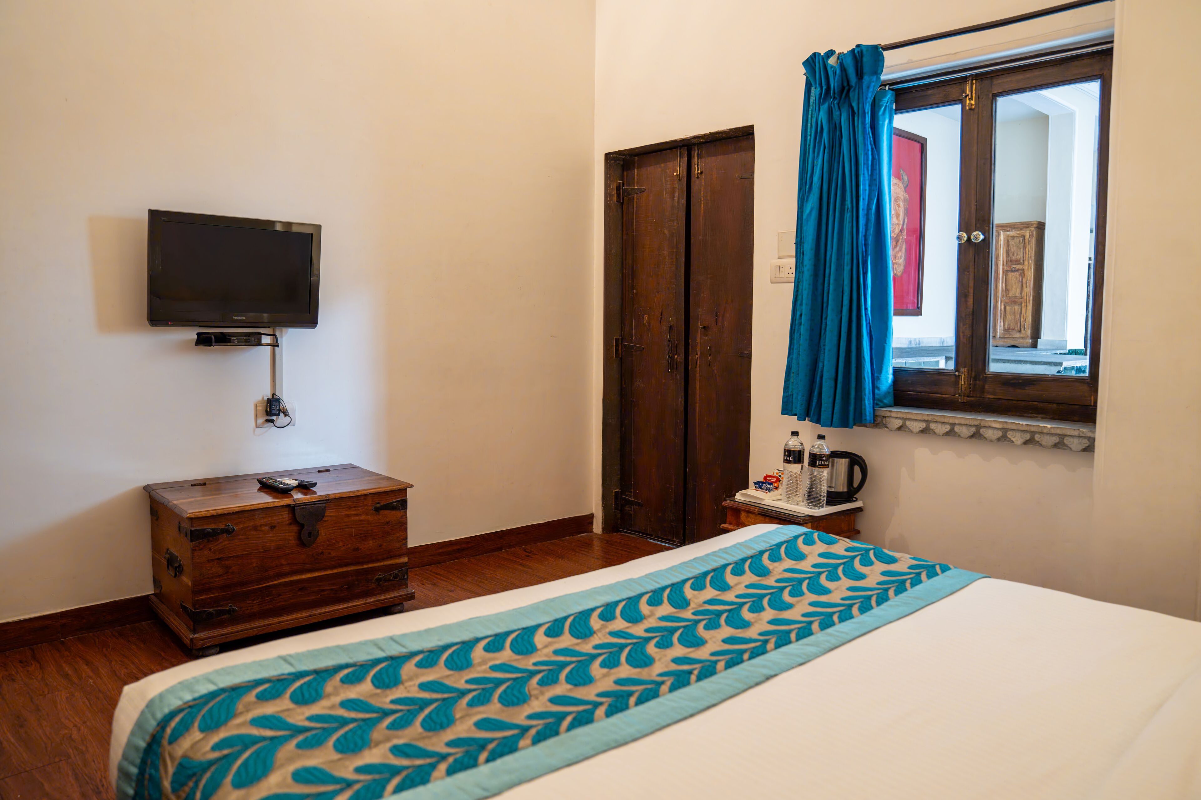 A bedroom with a bed, a wooden dresser, a television mounted on the wall, a window with curtains, and a door.