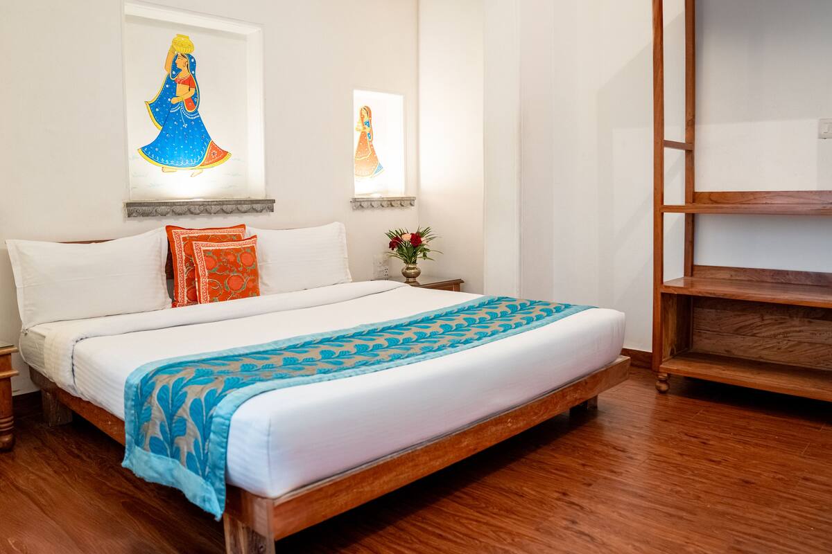 A bedroom with a wooden bed, white bedding, a blue and orange patterned throw, and two framed pictures on the wall.