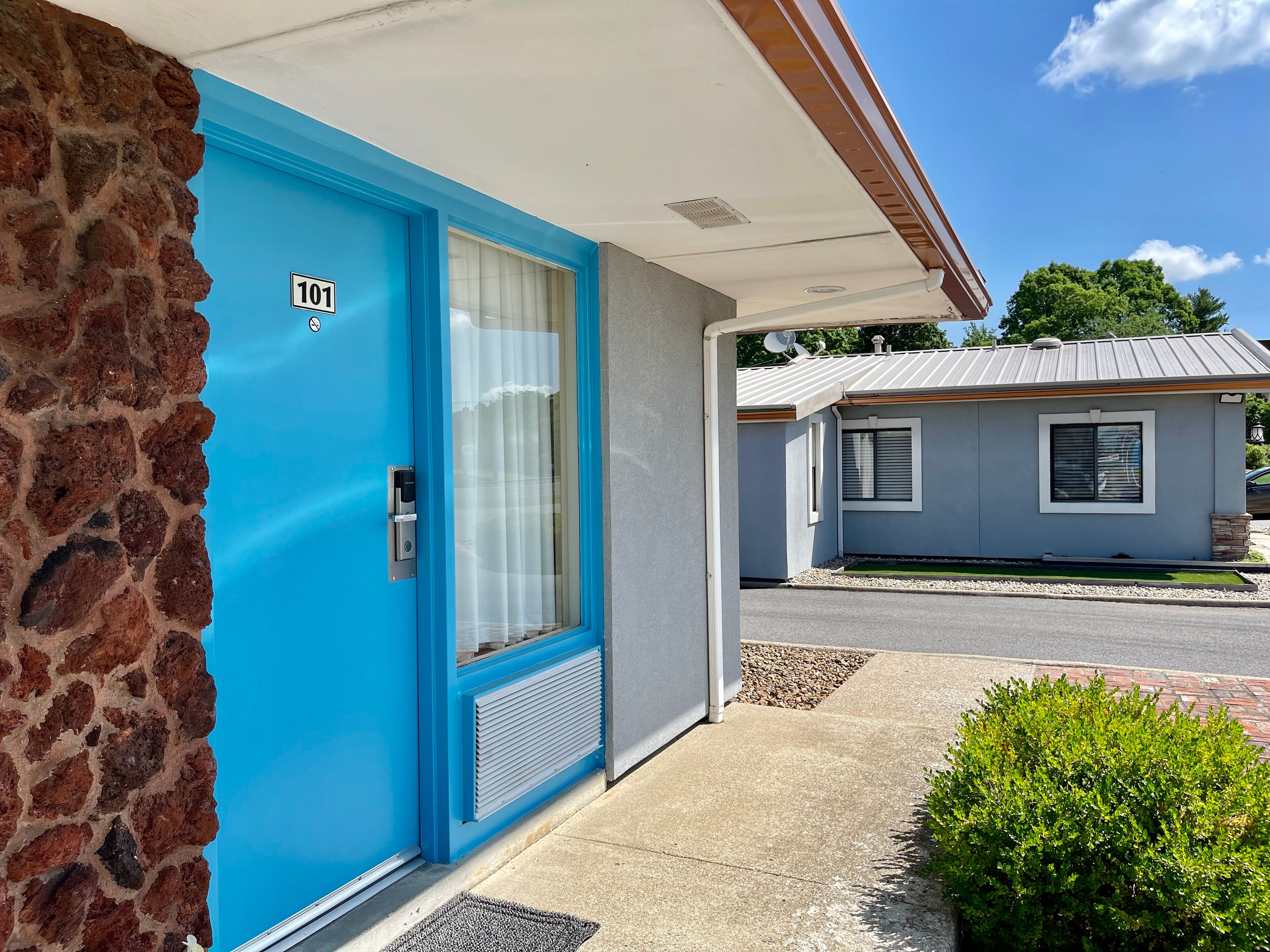 Ein Motel mit blauer Tür, auf der '101' steht, einer Steinmauer und einem kleinen Garten.