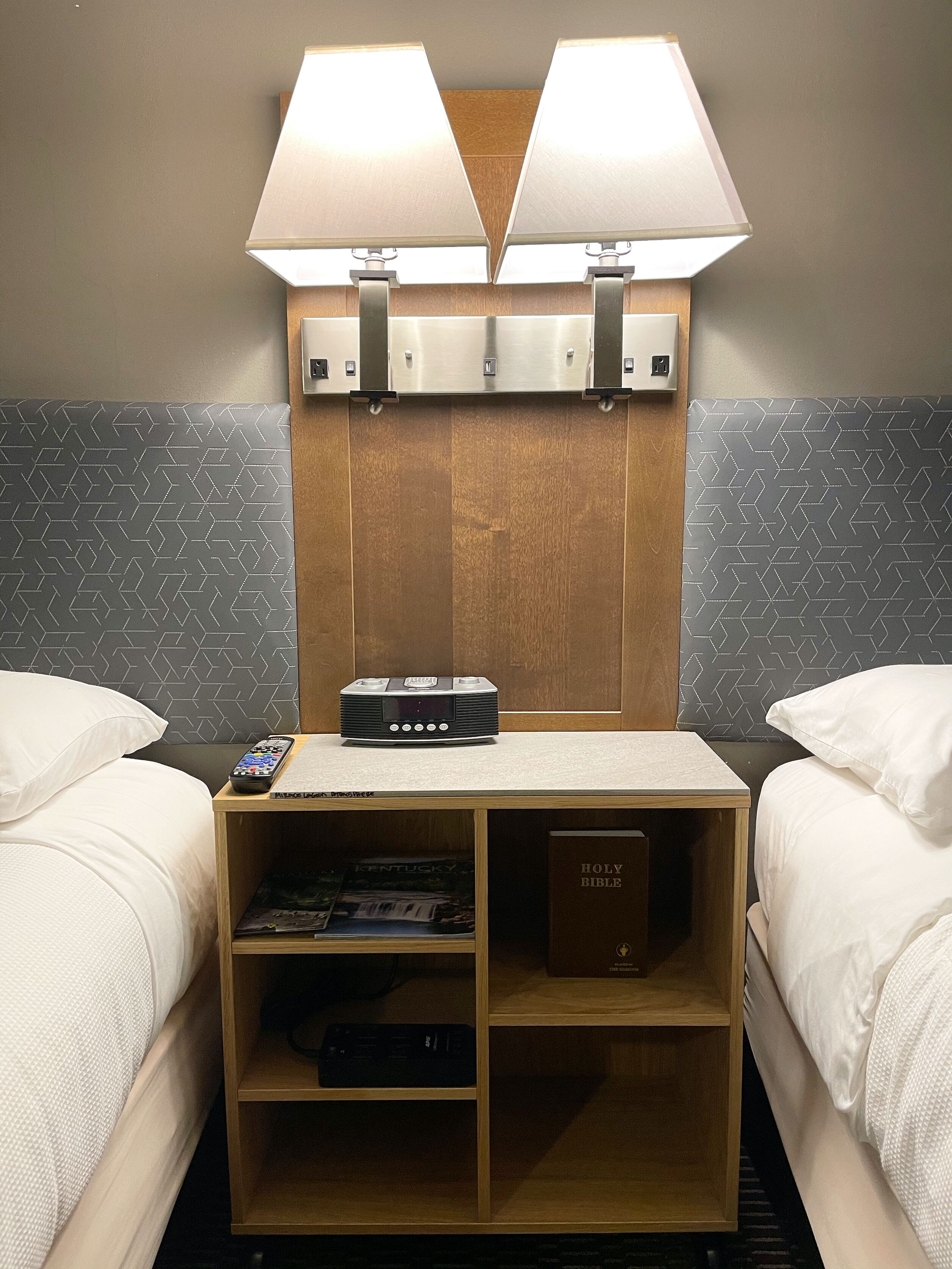 A hotel room with two beds, a wooden headboard, a nightstand with a lamp, a remote control, and a Bible.