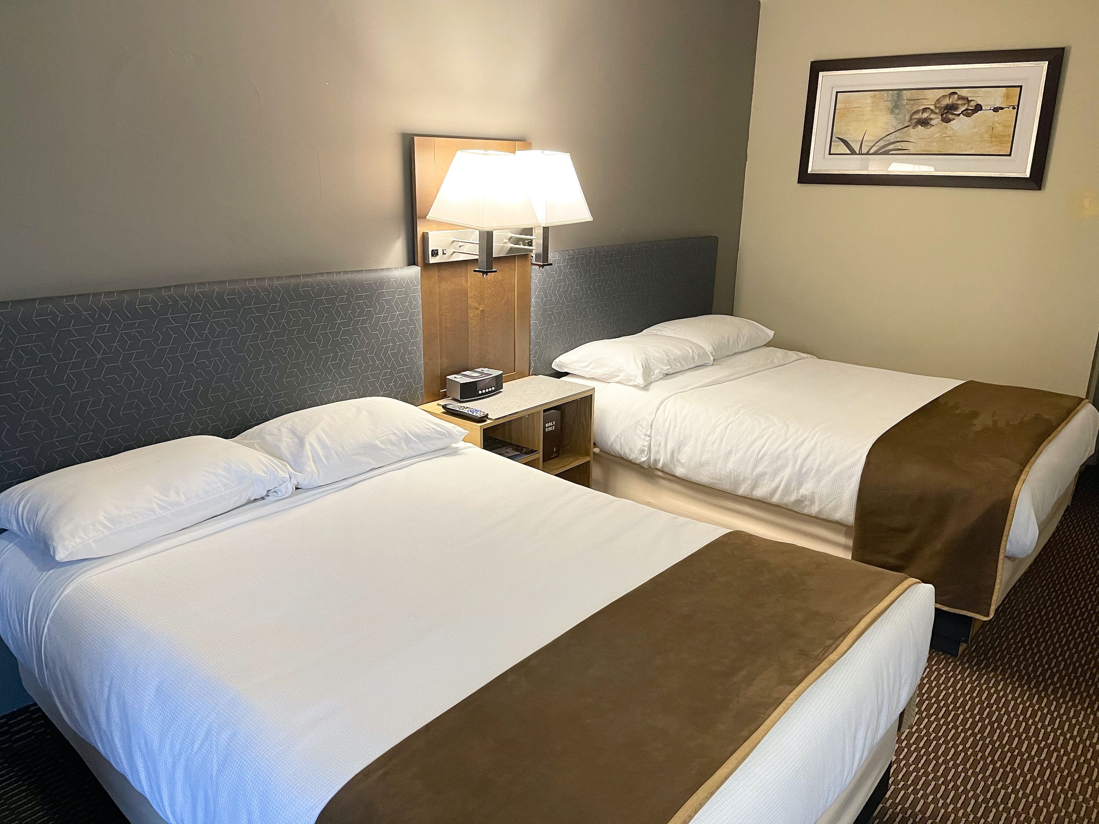 A hotel room with two beds, a nightstand with a lamp, a remote control, and a framed picture on the wall.