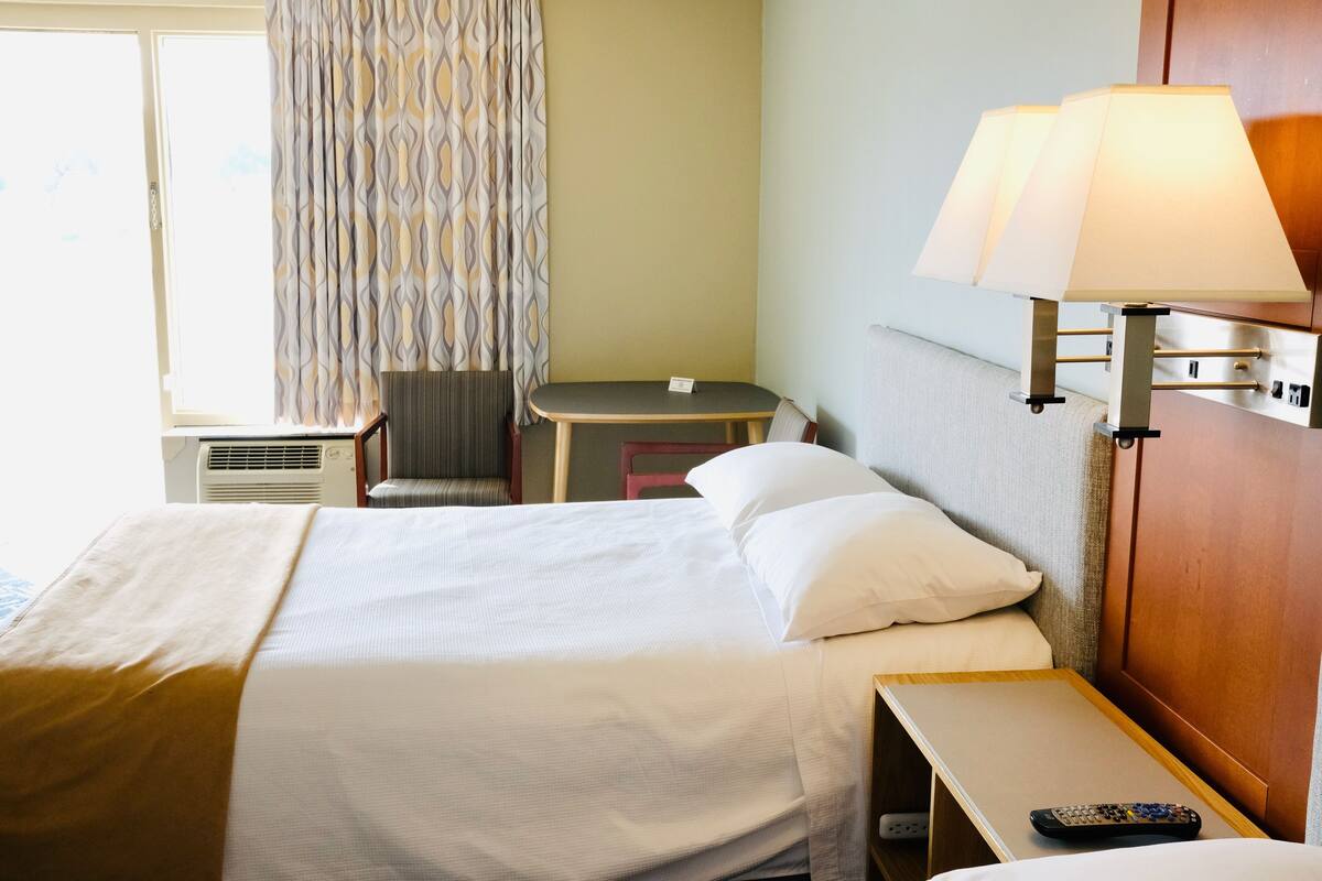 A hotel room with a bed, bedside table, lamp, chair, and a small desk with a chair.
