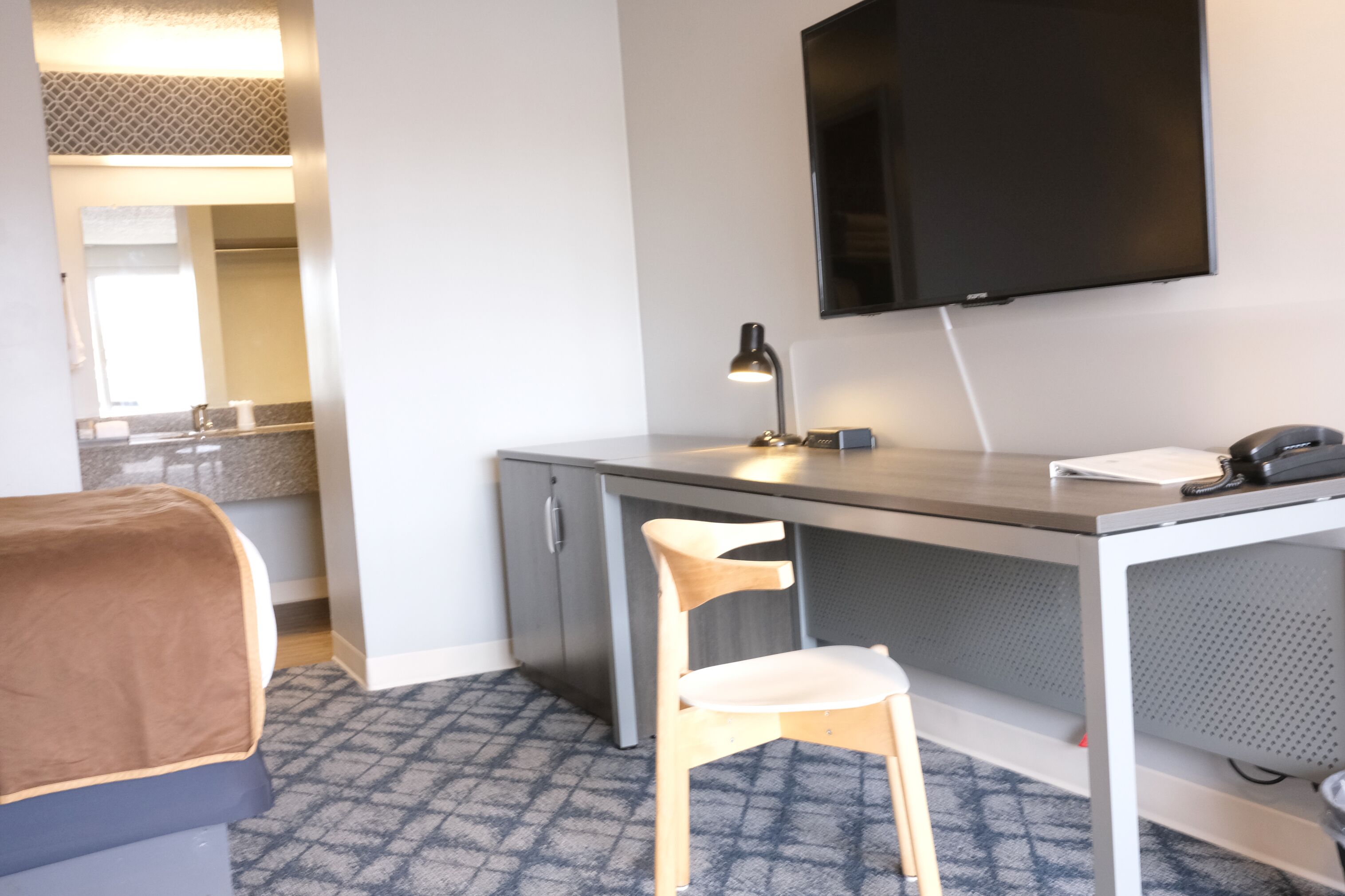 A hotel room with a bed, a desk with a chair, a television, and a bathroom visible in the background.
