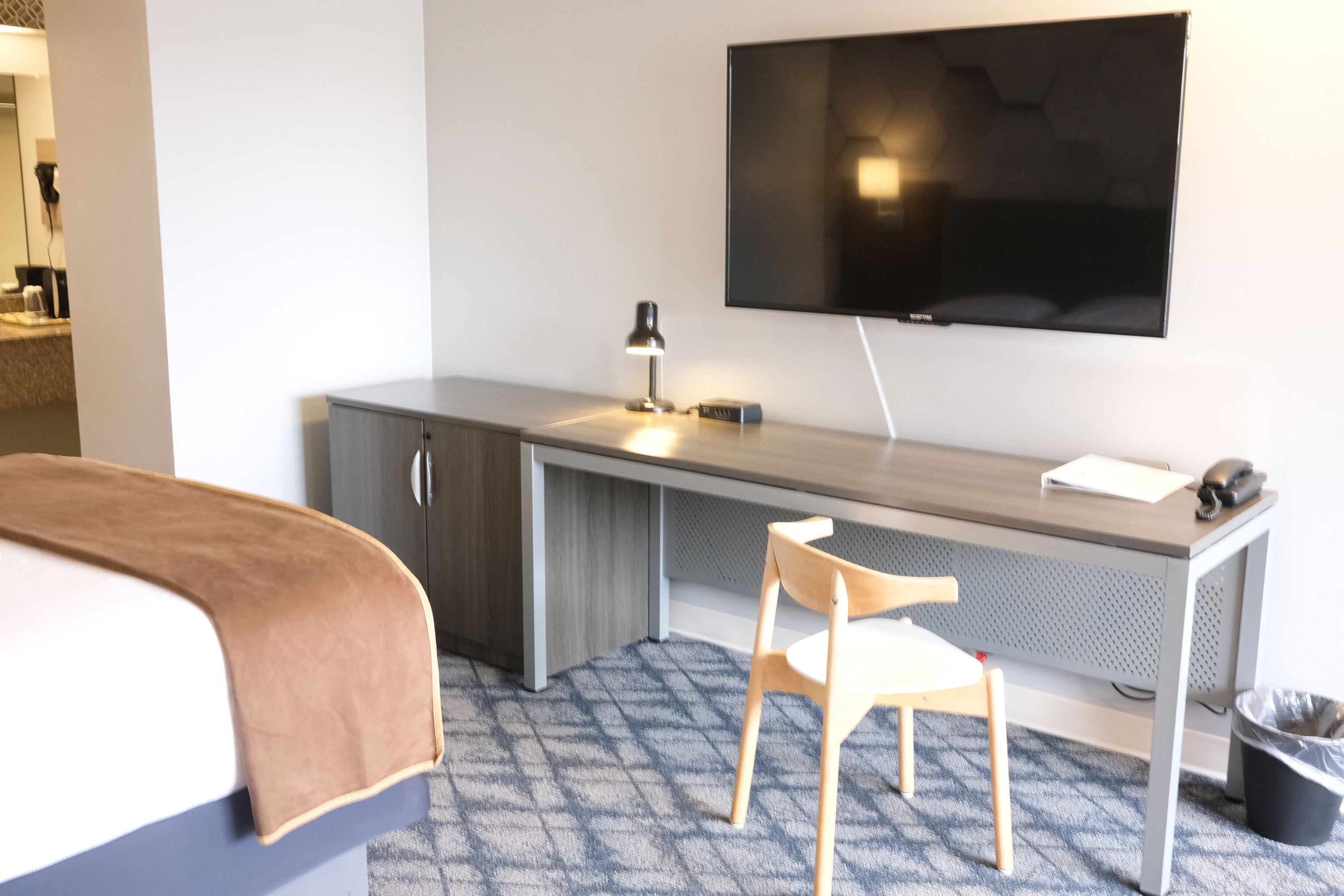 A hotel room with a bed, a desk with a chair, a television, and a trash bin.