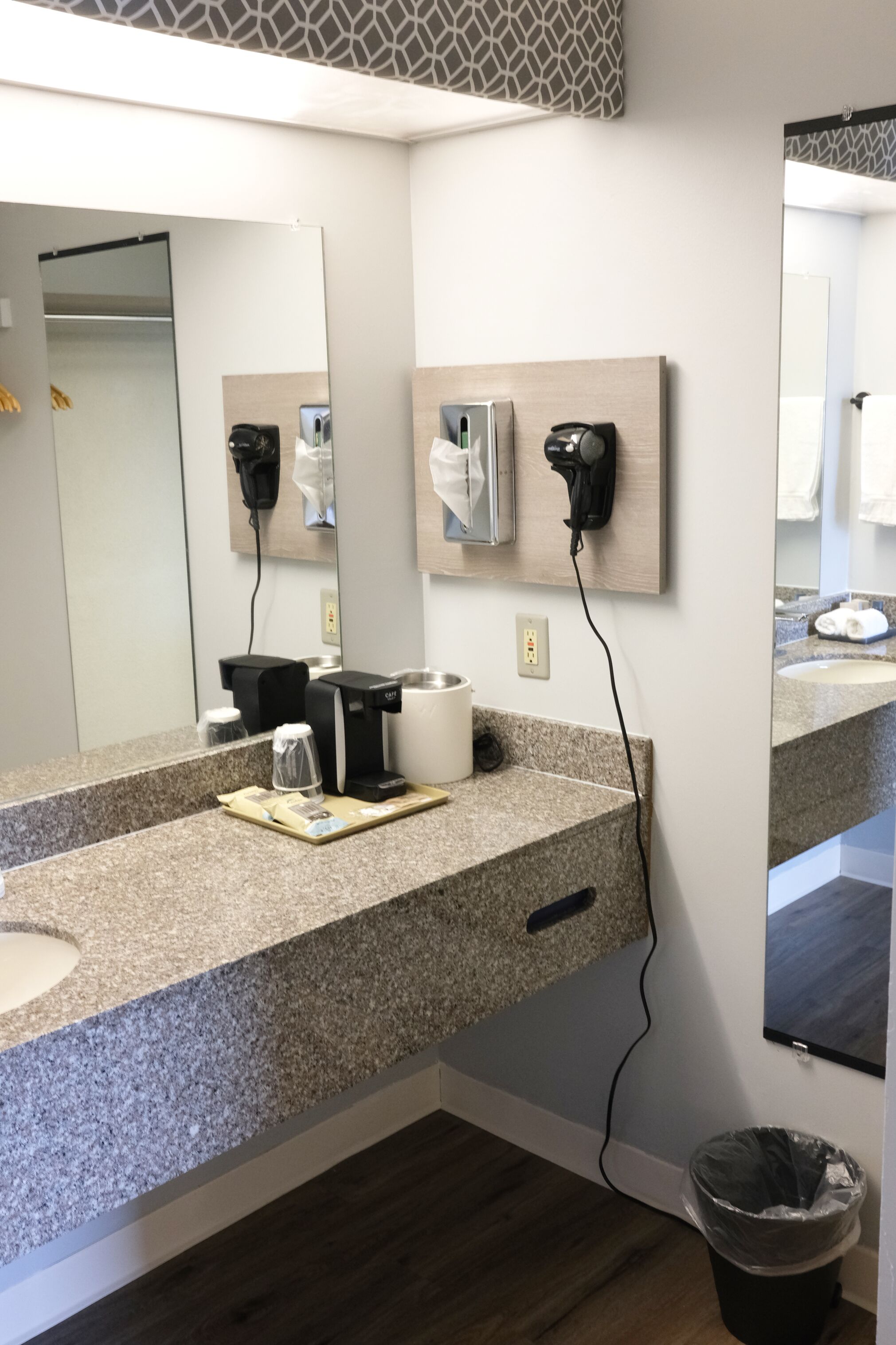 A hotel bathroom with a granite countertop, a sink, a mirror, a hairdryer, and a trash can.