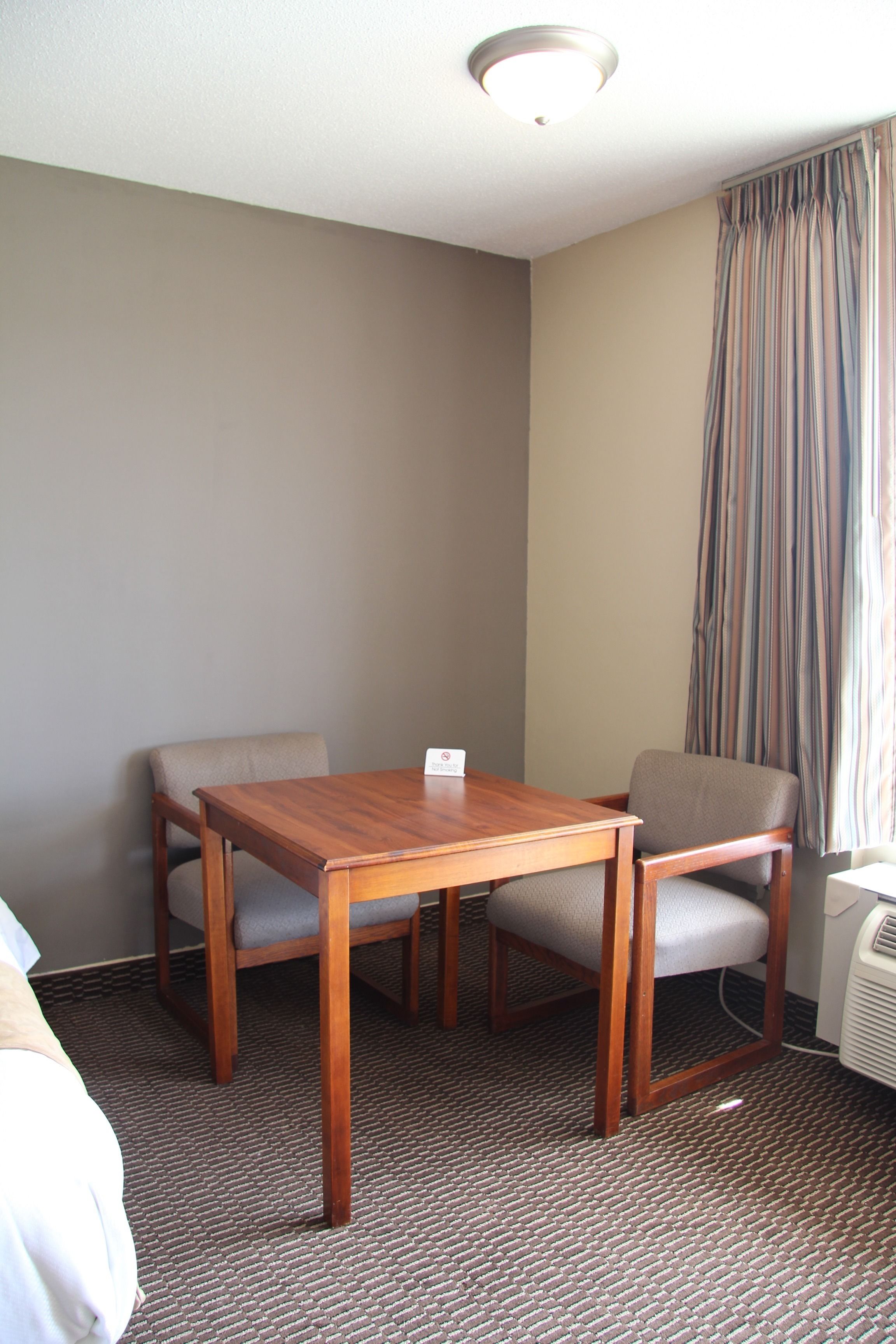 Habitación de hotel con una mesa de madera y dos sillas, una cama, una ventana con cortinas y un aire acondicionado instalado en la pared.