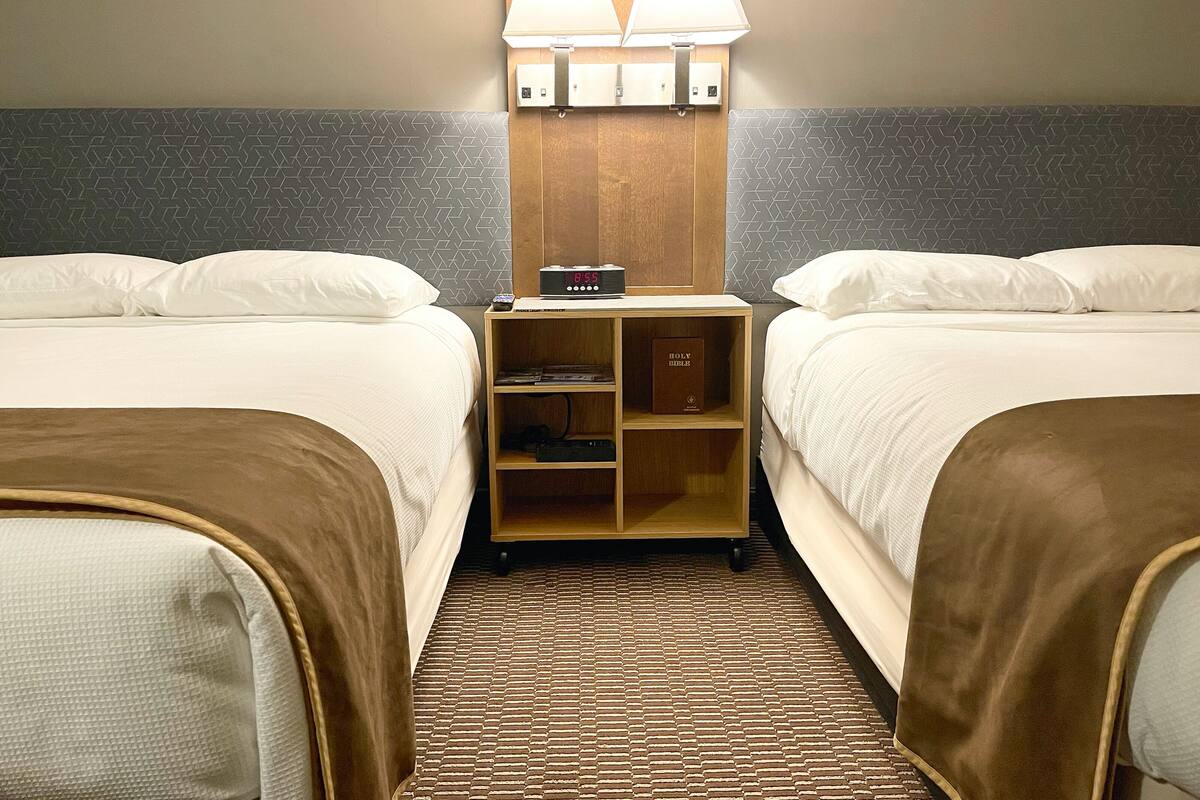 A hotel room with two beds, a nightstand with a lamp, a clock, and a Bible.