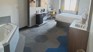Habitación de hotel con cama, escritorio, silla, kitchenette pequeña y baño.