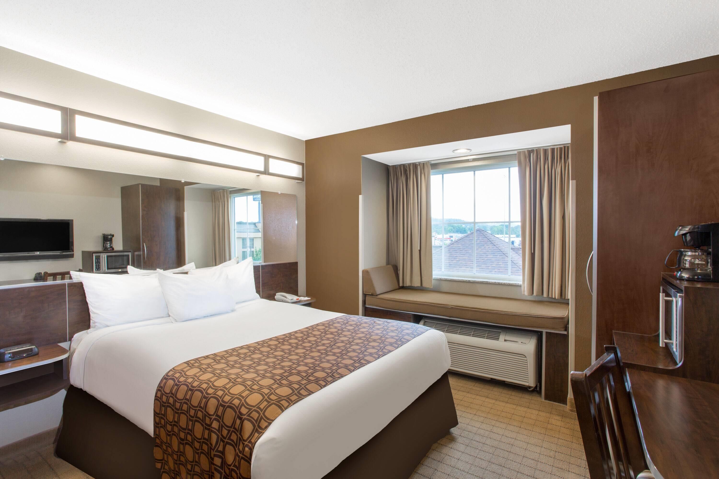 A hotel room with a large bed, a flat-screen TV, a desk with a telephone, a window with a view of the city, and a kitchenette area.
