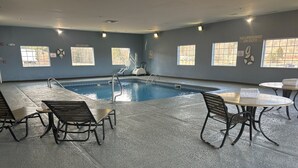 An indoor swimming pool area with poolside chairs and tables, a lifeguard station, and windows with safety signs. - Microtel Inn & Suites by Wyndham Mansfield (Mansfield)