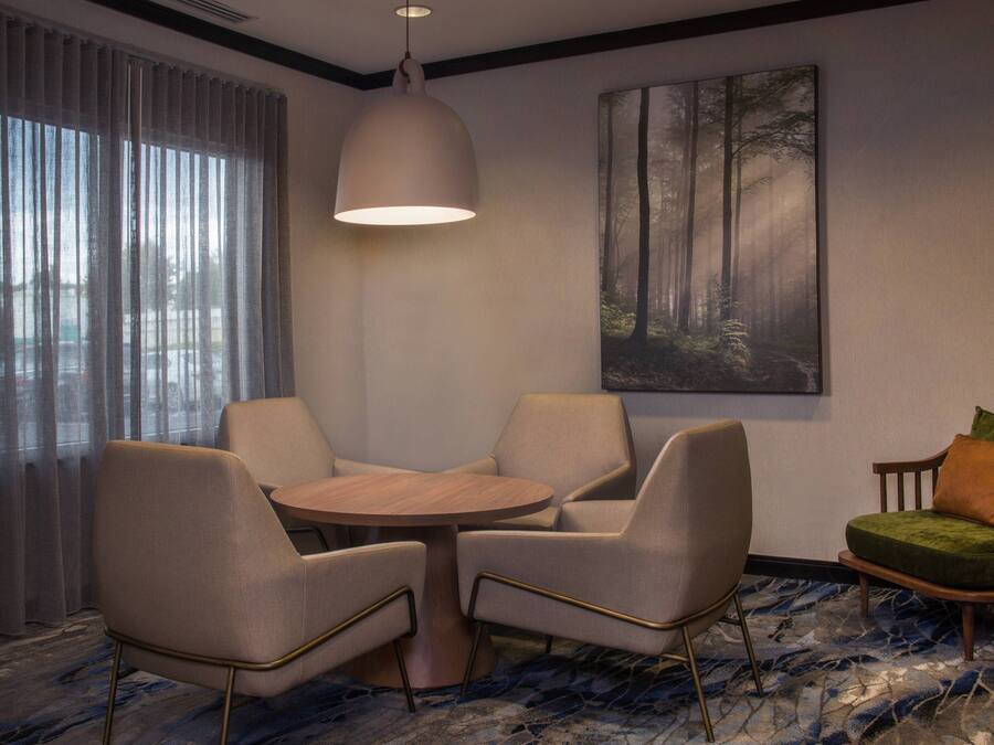 Una habitación moderna con una mesa redonda, cuatro sillas, una lámpara colgante, un cuadro de gran tamaño con temática forestal y vistas a través de cortinas traslúcidas.