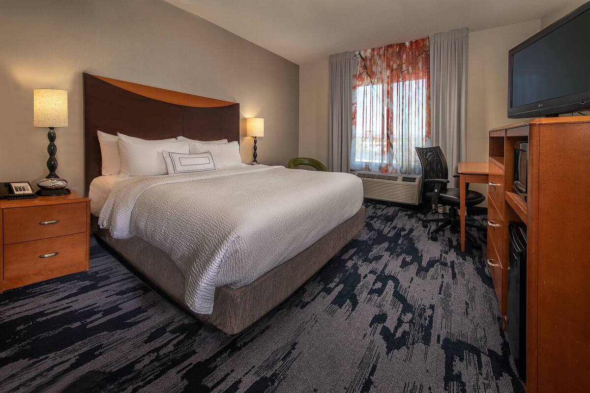 Ein Hotelzimmer mit einem großen Bett, zwei Nachttischen inklusive Lampen, einem Fernseher, einem Schreibtisch mit Stuhl und einem Fenster mit Vorhängen.
