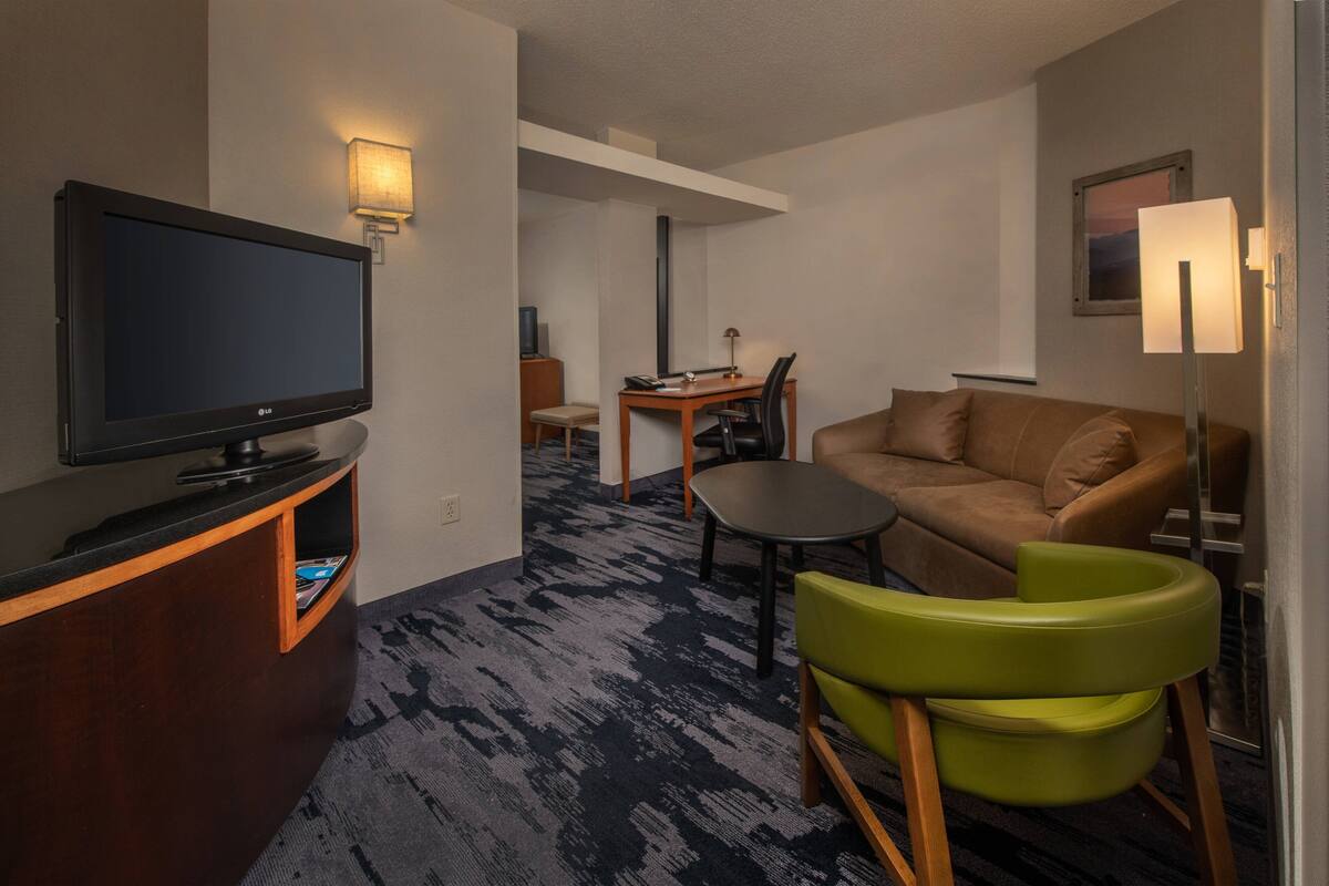 Ein Hotelzimmer mit Flachbildfernseher, einer Couch, einem kleinen Tisch, einem grünen Sessel und einer Lampe.